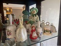Porcelain and ceramic decorative bells on glass shelf, various styles including floral and figurine bells.