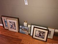 Multiple framed art prints and paintings arranged on hardwood floor against wall; some leaning.