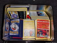 Open metal tin showing large stacks of Pokemon trading cards with colorful fronts and backs, including energy cards and character cards.