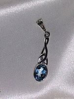 Pendant against white satin fabric showing detailed silverwork and faceted blue topaz oval gemstone.