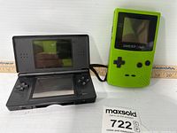 Two handheld consoles on a white surface: black Nintendo DS Lite on left and green Nintendo Game Boy Color on right.