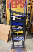 Full view of black and blue appliance dolly with straps leaning against a Pepsi sign in warehouse setting.