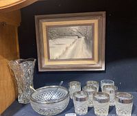 Framed winter landscape painting above cut glass bowl with silver rim, serving spoons, cut glass vase, and nine glasses on dark surface