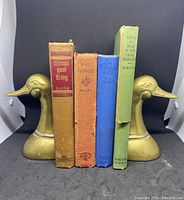 Front view of a pair of brass duck bookends holding four hardcover books with aged covers in red, orange, blue, and green.