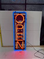 Brightly lit vertical OPEN sign with blue border and red neon letters, remote attached, cord visible.