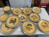 Large grouping of 22 ceramic plates laid out on a table, each plate featuring folk art style designs such as birds, animals, and geometric patterns, with yellow centers and speckled blue-green rims.