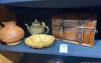 Photo showing signed ceramic teapot, pottery vase, yellow dish, and leather storage chest together on a shelf