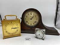 Three vintage mantle clocks shown together: brass Hamilton carriage clock on the left, wooden Sessions clock in the middle, and small clear acrylic WERItime clock on the right.