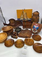 Photo shows the entire lot of vintage wooden decor items arranged on a white table with a yellow board in the background including bowls, plates, a wooden clock, ashtray, segmented dish, mounted plaque, model sailboat, and wooden canteen.