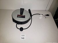 Brand-new round extension wrap cord with retractable cable and power plug, black and gray in color, shown on white surface.