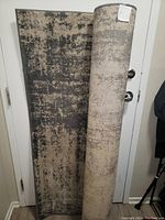 Photo of the rolled and partially unrolled floor rug showing abstract beige, gray, and black distressed pattern and dark edge binding.