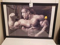 Framed black and white photograph of a muscular man flexing his right arm, with a visible lamp reflection on the glass