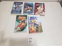 Photo showing five Disney animated VHS movie tapes on a white table with a lot number card.