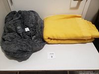 A photo showing the yellow fleece blanket and the folded plush grey heated blanket on a white table.