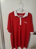 Red short sleeve polo shirt hanging on white hanger, showing front with white collar and Coca-Cola logo on left chest.