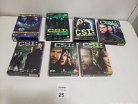 Photo showing all seven DVD box sets of CSI seasons 1 to 7 arranged in two rows on a white surface.