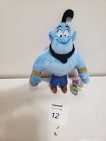 Front view of blue Disney Genie plush with cheerful facial expression, gold wrist cuffs, red waistband, and purple pants.