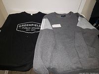 Front view of black men's T-shirt showing 'Greenfield Public House Est. 1923' text and gray sweater with white shoulder stripes.