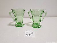 Photo showing a pair of light green glass milk and sugar bowls with pedestal bases and angular handles, placed side by side on a plain white surface.