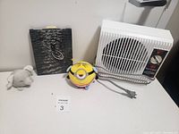 Group photo showing a white electric heater, a yellow Minion plush toy, a small grey plush toy, and a small framed picture with a boat.