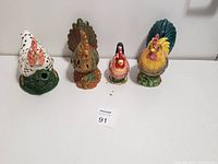 Four ceramic rooster and hen figurines arranged in a row on white surface against white wall.