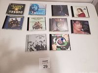 Ten various music CDs placed on a white table in two rows, showing album cover art and text.