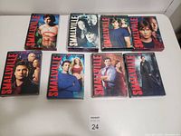 Nine DVD box sets of Smallville series seasons 1 to 9 arranged in two rows on a flat surface. Each case has different cover art featuring various characters from the series.
