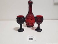 Front view of vintage red glass wine decanter with stopper and two matching embossed red glass wine glasses arranged side by side.