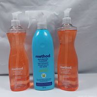 Three bottles: two orange translucent pump bottles with dish soap, one blue spray bottle with bathroom cleaner, all with Method branding.
