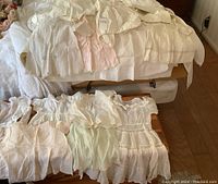 Photo of antique children's linen clothes laid out on tables, showing dresses and blouses in gentle pastel colors including white, light pink, and light green