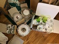 Overall image showing boxed Christmas china pieces including plates, bowls, mugs, and wrapped glass goblets on wooden floor