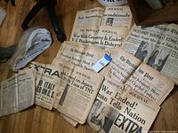 Photo of multiple historical newspapers laid out on a floor, showing headlines about WWII and Kennedy assassination