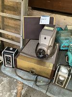 Photo showing two Kodak Duaflex cameras placed on and beside a vintage slide projector with original case.