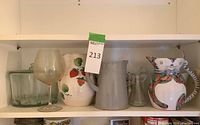 Shelf with the various pitchers, glassware including a wine glass, square glass container, white strawberry ceramic pitcher, grey ceramic pitcher, and a clear small pitcher.