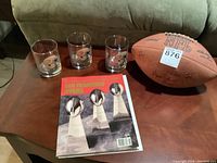 Photo showing three 49ers drinking glasses, an official NFL football with visible autographs, and a 49ers Super Bowl booklet on a wooden table.