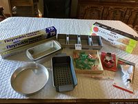 Photo showing the full lot including frost white tablecloth background with boxes of Reynolds freezer paper, a small round metal pan, three metal loaf pans including perforated and standard three-compartment pans, assorted plastic cookie cutters in a box, a metal snowman with heart cutter, and metal cake decorating tubes with tips.