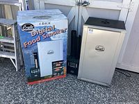 Box labeled "4 Rack Digital Food Smoker" next to assembled smoker tower and digital control unit with two black chimneys.
