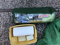 Coleman tent packed in green carry bag with label, yellow camp dish station containing soap holder, sponge, and soap bar.