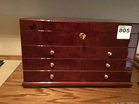 Closed wooden humidor with glossy reddish-brown finish and lock on the top drawer, with four drawers and gold-colored knobs.