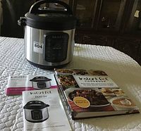 Photo shows Instant Pot Duo SV multicooker, two instruction manuals, and an Instant Pot cookbook arranged on a white quilted surface.