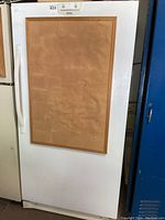 White upright freezer with a large cork bulletin board screwed onto the door front. The appliance is standing beside other items in a garage or storage area.
