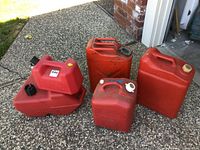 Photo showing assortment of five red plastic gas cans and one large red metal gas can on concrete pavement near garage.