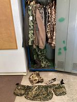 Photo showing multiple camouflage jackets hanging inside a locker alongside camouflage pants and other apparel items. A camouflage utility belt with pouches is placed on the floor in front of the locker.