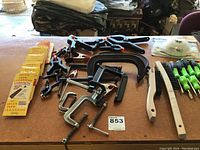 Top view of table with assorted clamps, pipe cleaners in yellow packaging, screwdrivers with green handles, and wire brushes arranged on a table surface