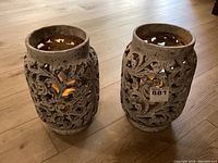 Front angled view of the two resin decorative carved pots on wooden floor, showing detailed floral cut-out pattern and weathered grey finish.