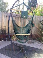 Full view of the green hanging chair suspended from a heavy black metal frame with curved base, wooden support dowels, small seat cushion, and footrest.