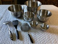 Six stainless steel mixing bowls of various sizes displayed with three kitchen utensils on a tablecloth.
