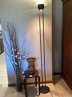 Standing metal floor lamp beside small round wooden table holding a large decorative ceramic vase. A tall clear glass vase with floral branches next to the lamp.