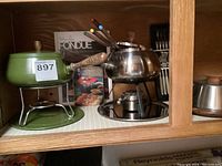 Two vintage fondue pots with cookbooks, fondue forks inside stainless pot, and cheese server displayed on wooden shelving