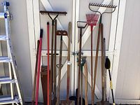 Photo showing a variety of garden tools including rakes, shovels, axes, and broom leaning against a white door.